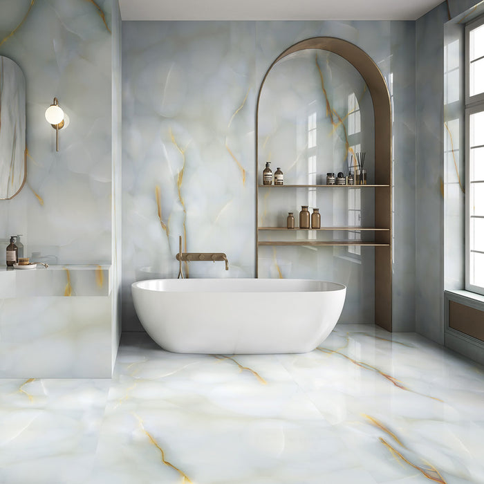 Bathroom interior featuring Fantasia Turchese Blue Onyx Polished 60 × 120cm stone effect wall and floor tiles, showcasing soft blue-grey tones with flowing gold veining, paired with a freestanding bath, brushed brass fittings, and an arched shelving niche for a calm, spa-like finish.
