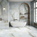 Bathroom interior featuring Fantasia Turchese Blue Onyx Polished 60 × 120cm stone effect wall and floor tiles, showcasing soft blue-grey tones with flowing gold veining, paired with a freestanding bath, brushed brass fittings, and an arched shelving niche for a calm, spa-like finish.
