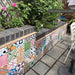 Outdoor garden featuring Fihrdos Mini Patchwork 15cm x 15cm Moroccan style tiles across a raised planter, combining colourful geometric and floral patterns with dark brick edging, paving and lush greenery.