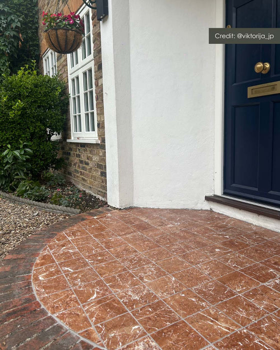 Outdoor entrance featuring Fitzrovia Rosso 15.2cm x 15.2cm natural marble floor tiles with a tumbled finish, laid beside traditional brickwork, a navy front door and surrounding greenery.