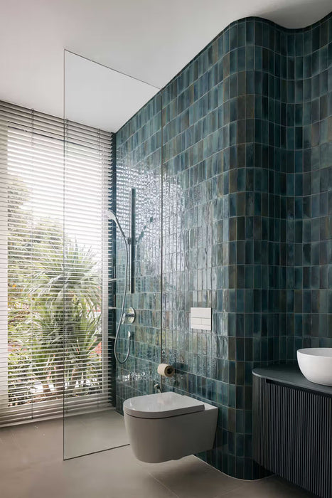Modern bathroom featuring Fusion Marine Blue 6.5x20cm gloss brick wall tiles, showcasing rich blue-green shade variation wrapped around curved walls, paired with a frameless walk-in shower, dark fluted vanity, white basin and large window for a contemporary spa-inspired finish.