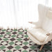 Living room setting featuring Geometry Angles Patterned Tile in 20cm x 20cm, arranged in a repeating geometric pattern with green, black, and off-white triangular shapes. A soft white armchair sits on the tiled floor beside sheer white curtains, creating a bright and airy atmosphere.