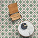 Top-down interior view featuring Geometry Green Clover 20x20cm patterned floor tiles in green, white and soft red tones, styled with a woven chair and marble table in a relaxed, contemporary living space.