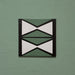 Single tile image showing the Geometry Hourglass 20x20cm patterned tile with alternating green and cream hourglass shapes outlined in charcoal, photographed against a textured sage-green background.