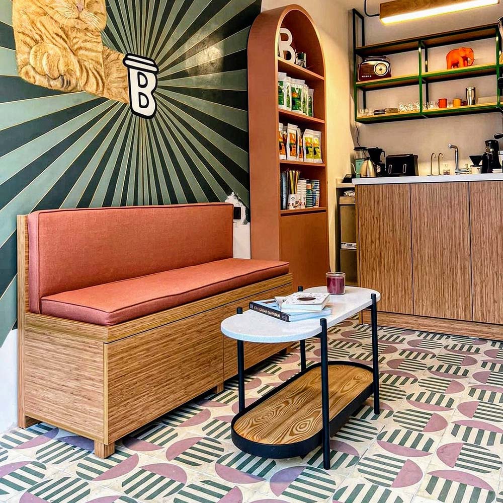 Café setting featuring Geometry Seritina 20x20cm patterned floor tiles with green striped and pink semicircle motifs on a cream base, styled with a wooden bench, terracotta-toned cushion, modern shelving, and a marble-topped coffee table.