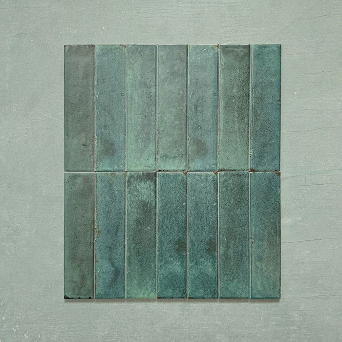 Group of Izmir Brick Green Gloss 6x25cm ceramic tiles displayed on a soft green background, highlighting the deep emerald tones, subtle shade variation, and glossy handcrafted effect finish.