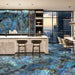 Open-plan kitchen and dining area featuring Illuminati Precious Stone Labradorite 60 × 120cm polished porcelain floor tiles, showcasing vivid blue gemstone-inspired veining, paired with marble-effect cabinetry, dark shelving, and contemporary furnishings.