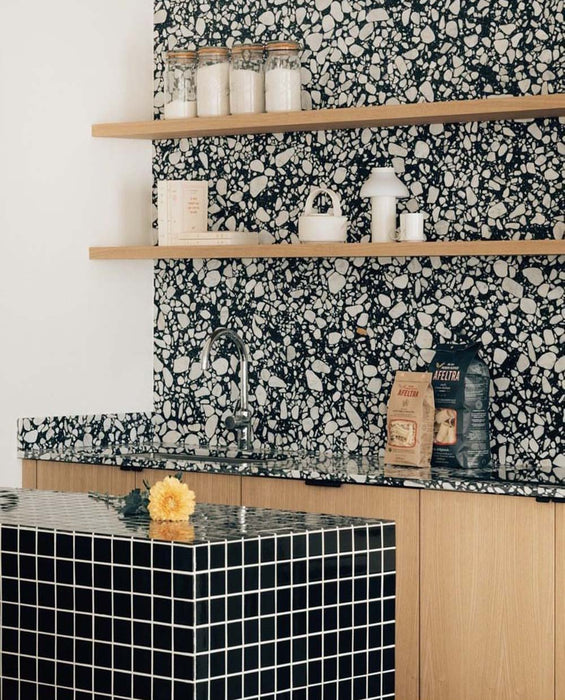 Kitchen setting featuring Noventa Terrazzo Nero 59x118cm matt porcelain wall and floor tiles, showcasing a bold terrazzo effect with contrasting white stone fragments set against a deep black base, used as a statement splashback and worktop detail, paired with warm wood cabinetry, open shelving and crisp black accents for a contemporary, design-led finish.