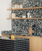 Kitchen setting featuring Noventa Terrazzo Nero 59x118cm matt porcelain wall and floor tiles, showcasing a bold terrazzo effect with contrasting white stone fragments set against a deep black base, used as a statement splashback and worktop detail, paired with warm wood cabinetry, open shelving and crisp black accents for a contemporary, design-led finish.