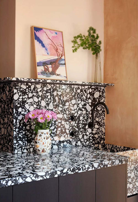 Bathroom vanity setting featuring Noventa Terrazzo Nero 59x59cm matt porcelain wall and floor tiles, showcasing a bold terrazzo effect with striking white stone fragments set against a deep black base, wrapped seamlessly across surfaces and paired with warm plaster tones, soft lighting and matte black fittings for a refined, design-led finish.