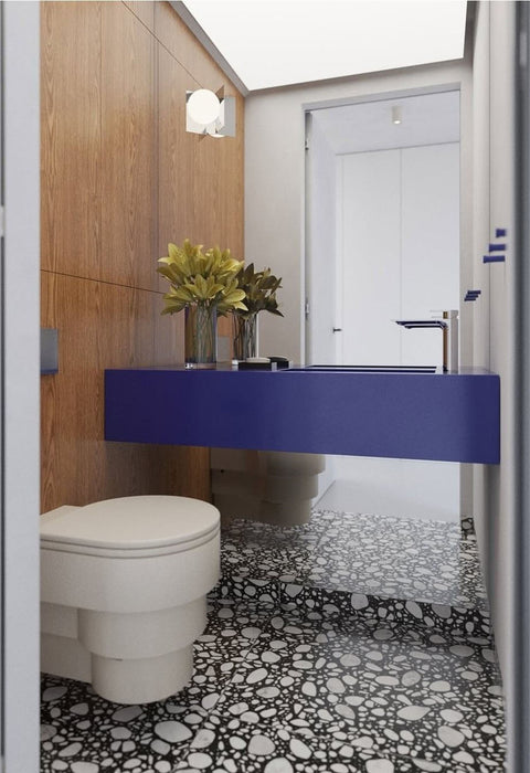 Bathroom setting featuring Noventa Terrazzo Nero 59x59cm matt porcelain wall and floor tiles, showcasing a bold terrazzo effect with crisp white stone fragments set against a deep black base, carried across both floor and stepped detailing, paired with warm wood panelling and a striking cobalt vanity for a contemporary, design-led finish.
