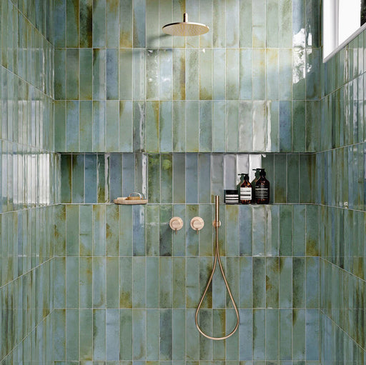 Contemporary shower featuring Izmir Brick Celadon Green Gloss 6x25cm ceramic wall tiles, styled with brushed brass fixtures, built-in shelving with amber bottles, and natural light reflecting on the glossy surface.