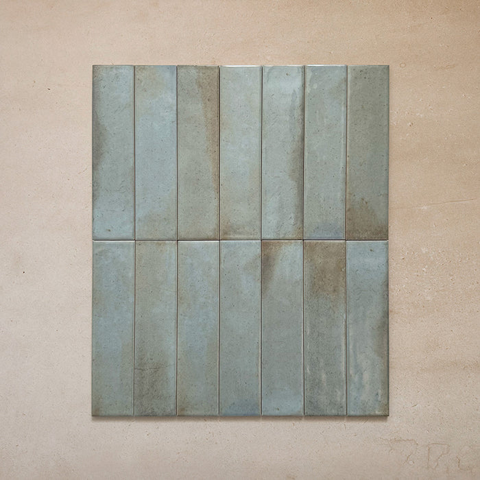 Group of Izmir Brick Celadon Green Gloss 6x25cm ceramic tiles displayed on a beige surface, showing subtle colour variation, glossy glaze, and handcrafted texture.