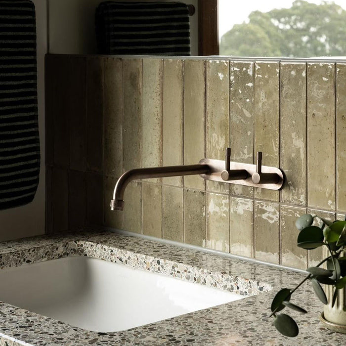 Bathroom featuring Izmir Sage Green gloss brick tiles in a 6 × 25cm ceramic format, installed vertically to create a soft, elongated look, with subtle tonal variation and a high-gloss finish that reflects natural light, paired with warm wood panelling, terrazzo surfaces and minimalist brass fittings for a calm, spa-inspired interior.