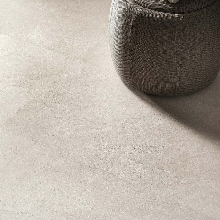 Close-up of Jeddah Avorio Neutra Stone 30x60cm stone-effect floor tiles, showcasing warm ivory and beige tones with delicate natural stone-inspired veining, paired with a soft upholstered pouffe in a contemporary interior.