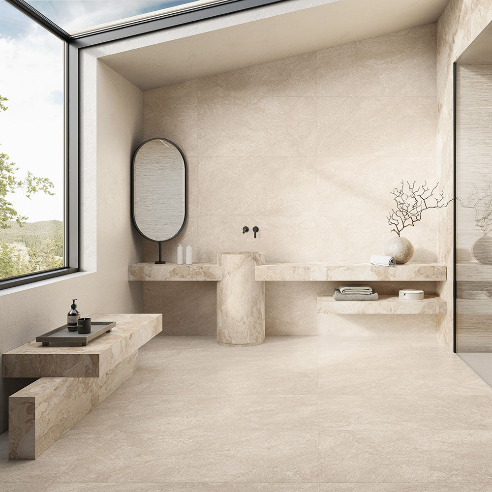 Contemporary bathroom featuring Jeddah Avorio Neutra Stone 60x60cm stone-effect wall and floor tiles, showcasing warm ivory and beige tones, paired with floating stone shelves, a pedestal basin, black fittings and minimalist décor.