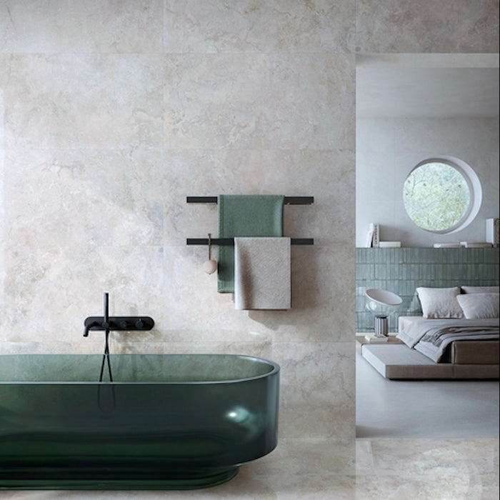 Bathroom featuring Jesi Travertine 60x60cm wall and floor tiles, showcasing soft travertine effect surfaces in warm cream and beige tones, paired with a translucent green freestanding bath, black brassware and sage green accents for a luxurious, spa-inspired interior.