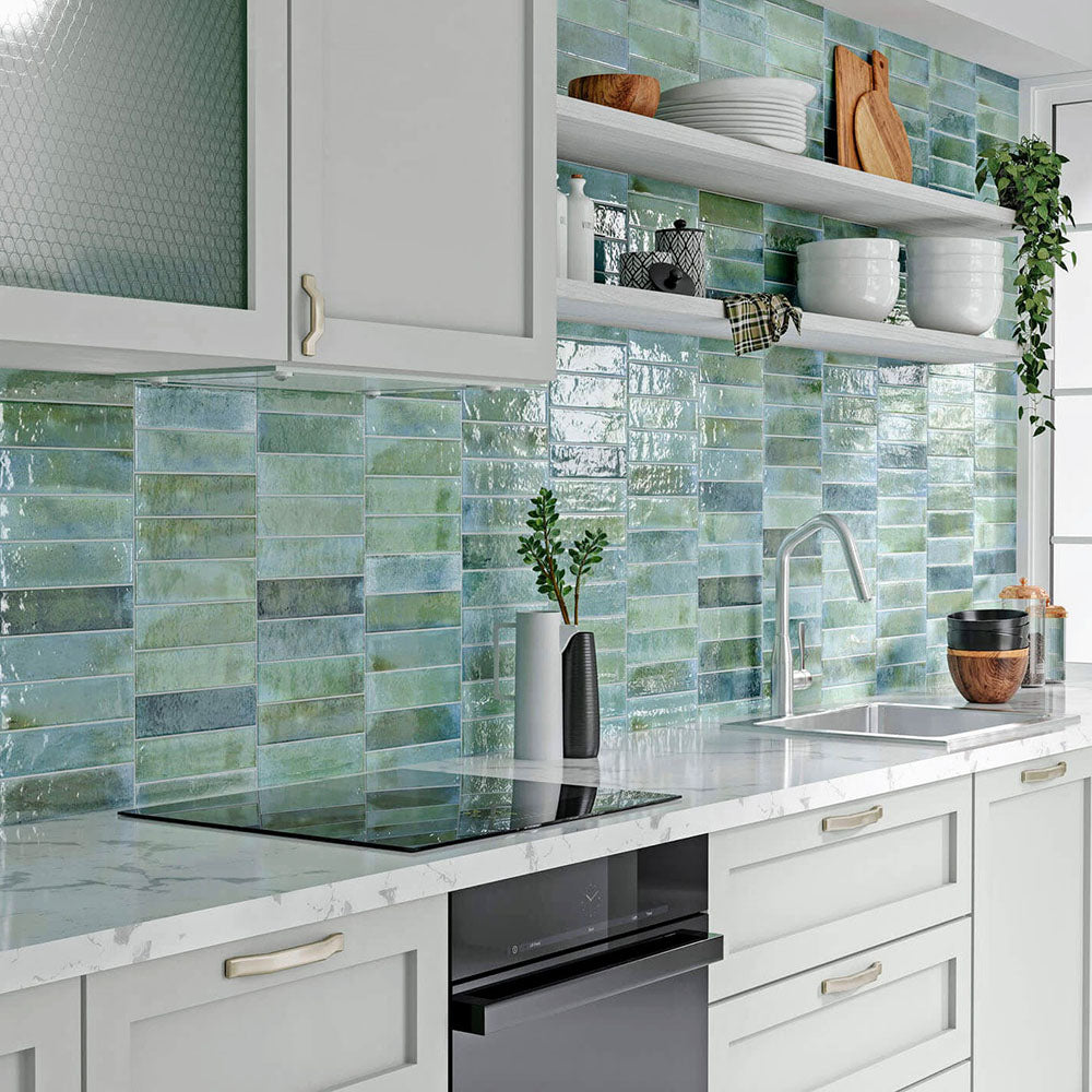 Modern kitchen featuring Izmir Brick Celadon Green Gloss 6x25cm ceramic wall tiles, styled with white shaker cabinets, marble-effect countertops, and open shelving with wooden boards, plants, and ceramic dishes.