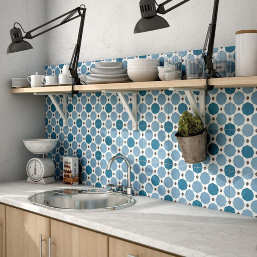 Kitchen setting featuring Carnaby Sapphire Victorian-style 20x20cm porcelain wall and floor tiles, used as a vibrant blue and white geometric backsplash, styled with light wood cabinetry, open shelving with tableware, industrial-style wall lamps, and a marble-look countertop.