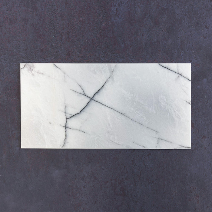 Fitzrovia Lilac 30.5x61cm natural marble tiles with a honed finish, featuring a soft white base with subtle lilac undertones and fine grey veining, shown as a single tile sample laid horizontally against a dark textured backdrop.