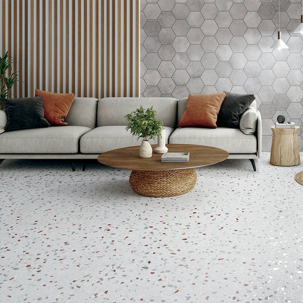 Living room featuring Confetti Colours 66×66cm terrazzo-style porcelain floor tiles with multicoloured chips on a white base, styled with a neutral sofa, wooden slat wall, grey hexagon feature wall, and a round woven coffee table for a modern, warm aesthetic.