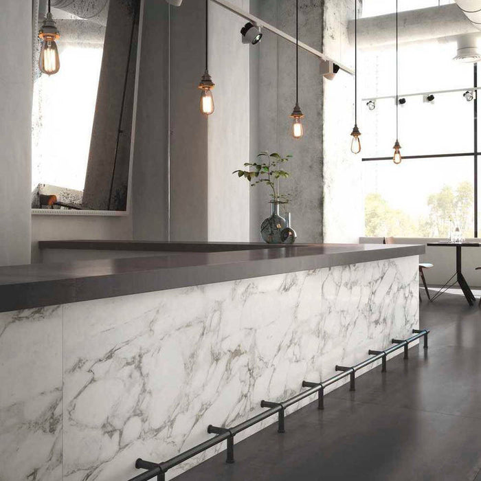 Bar and dining setting featuring Maine Calacatta Matt Marble 60 × 60cm porcelain wall tiles, styled across the counter front to showcase soft grey veining on a clean white base, paired with dark worktops, industrial lighting, and polished concrete for a refined, contemporary contrast.
