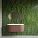 Bathroom interior featuring Malaga Tourmaline Green Brick 6.5 × 32.8cm gloss ceramic wall tiles, laid in a stacked brick pattern with rich green tonal variation and a reflective finish, paired with a sculpted wood vanity unit, brushed brass fittings, and an organic-shaped mirror for a refined, contemporary look.