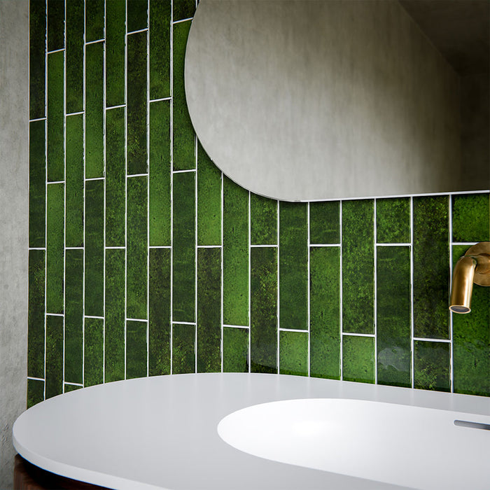 Bathroom interior featuring Malaga Tourmaline Green Brick 6.5 × 32.8cm gloss ceramic wall tiles, laid in a stacked brick pattern with rich green tonal variation and a reflective finish, paired with a sculpted wood vanity unit, brushed brass fittings, and an organic-shaped mirror for a refined, contemporary look.