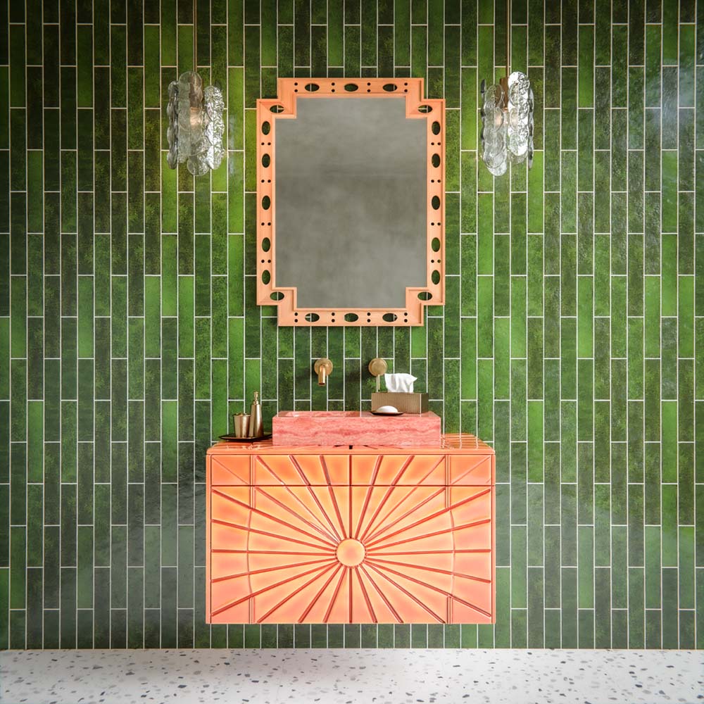 Bathroom feature wall showcasing Malaga Tourmaline Green Brick 6.5 × 32.8cm gloss ceramic wall tiles, laid in a vertical stacked pattern with deep green tonal variation and a glossy handcrafted finish, paired with a statement coral vanity unit, brushed brass fittings, and decorative lighting for a bold, design-led interior.