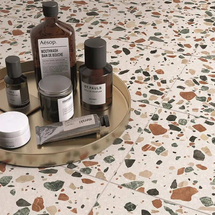 Flatlay featuring Dino Terrazzo Blanco Drops 20x20cm terrazzo-style ceramic floor tiles, styled with amber glass bottles and skincare products on a gold tray, highlighting the earthy terrazzo pattern.