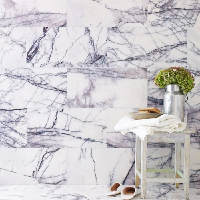 Interior setting featuring Fitzrovia Lilac 30.5 x 61cm honed natural marble tiles installed in a staggered brick layout, showcasing soft white tones with flowing lilac-grey veining, styled with a rustic stool, soft textiles, and decorative accents for a calm, considered finish.