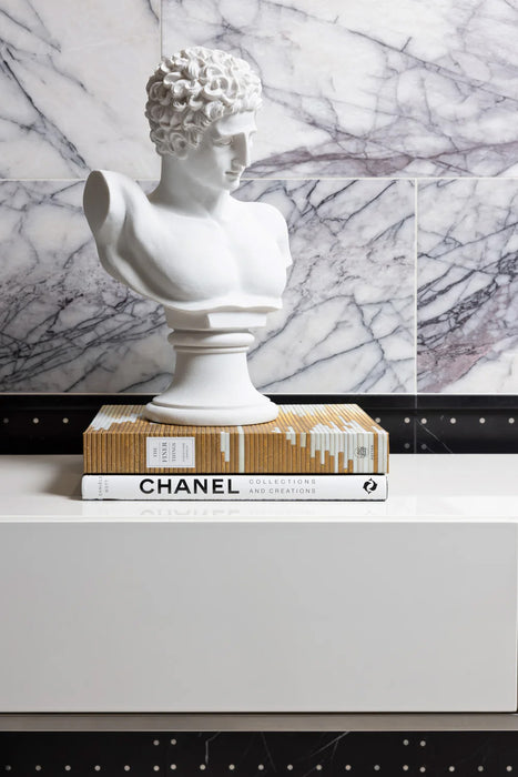 Interior detail featuring Fitzrovia Lilac 30.5 x 61cm honed natural marble tiles installed as a statement wall, showcasing a soft white base with expressive lilac-grey veining, styled with a sculptural bust and decorative books for a refined, gallery-inspired setting.