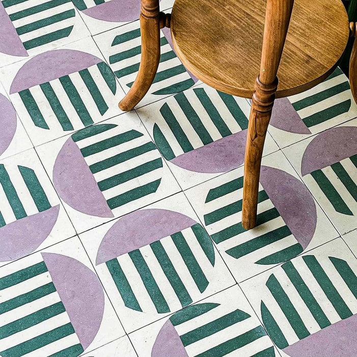 Marketing image featuring Geometry Seritina 20x20cm patterned floor tiles with green stripes and mauve semicircles on a cream background, shown beneath a wooden chair to highlight the bold repeating design.