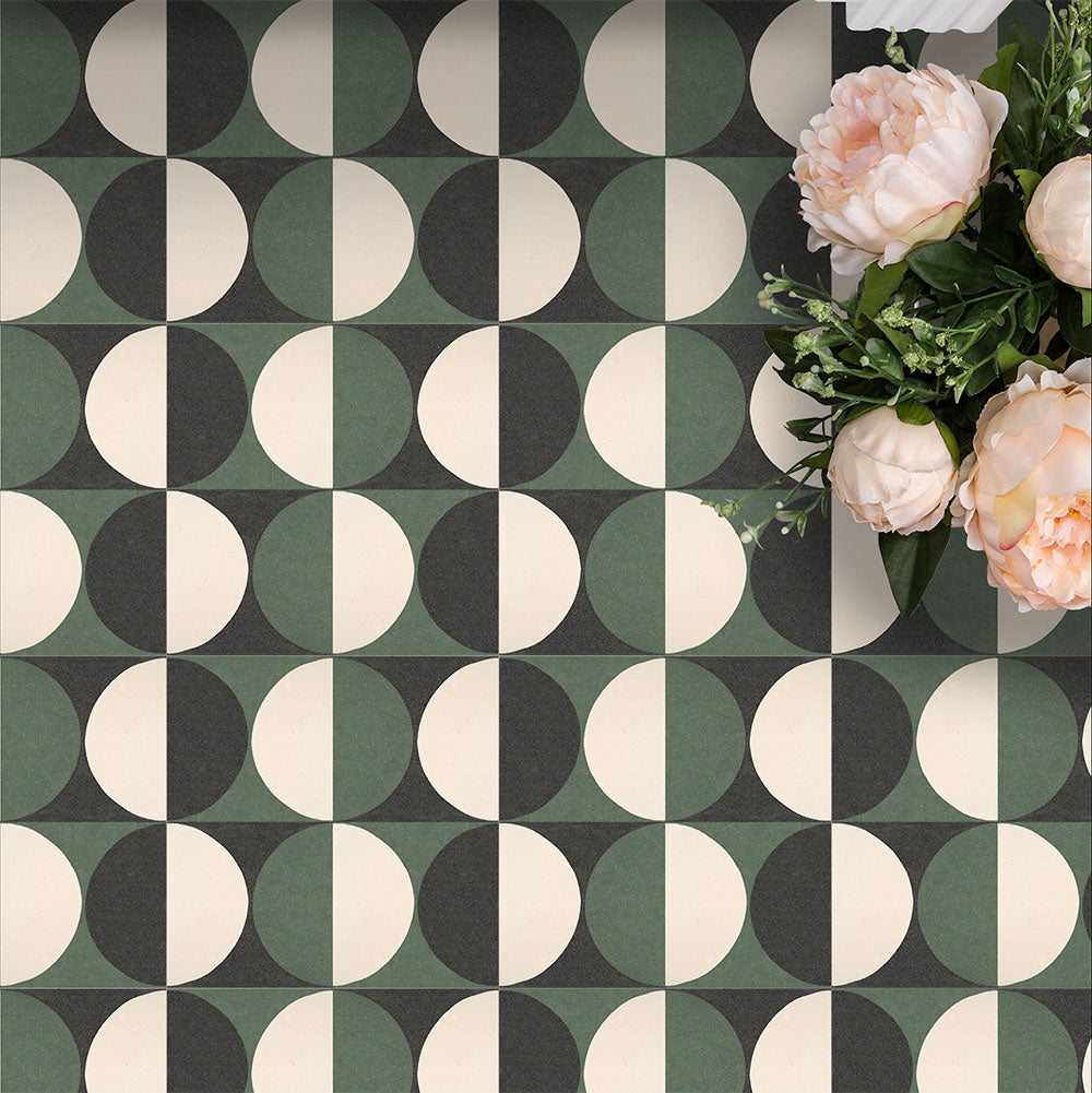 Marketing image featuring Geometry Circulo 20x20cm geometric patterned floor tiles in green, black, and cream tones, styled with soft pink peonies arranged in the corner for a decorative flatlay effect.