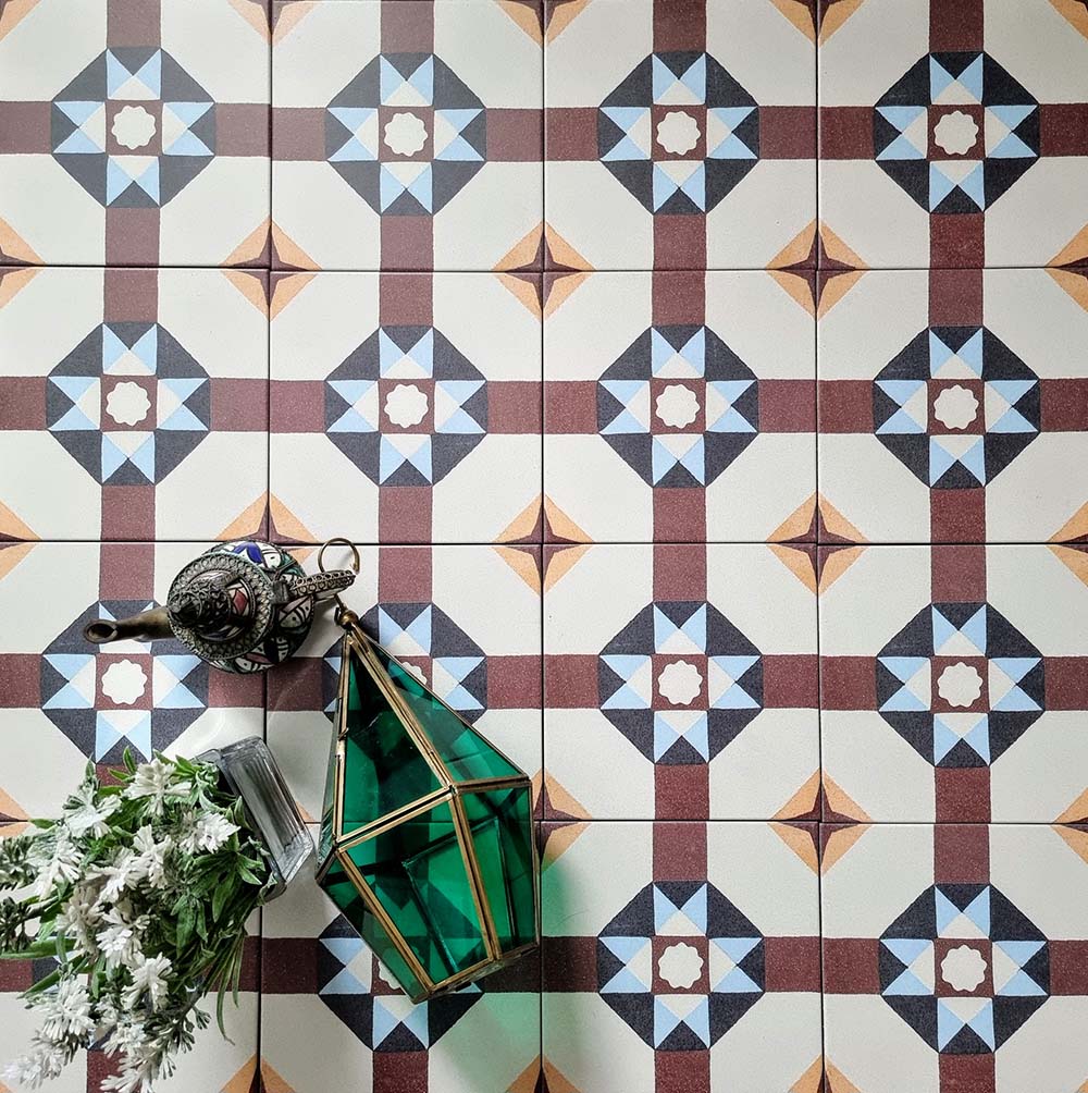 Overhead view of Europa Berlin 20x20cm Victorian Moroccan style floor tiles in burgundy, blue, mustard and cream tones, forming a geometric repeating pattern, styled with decorative lantern and floral arrangement.