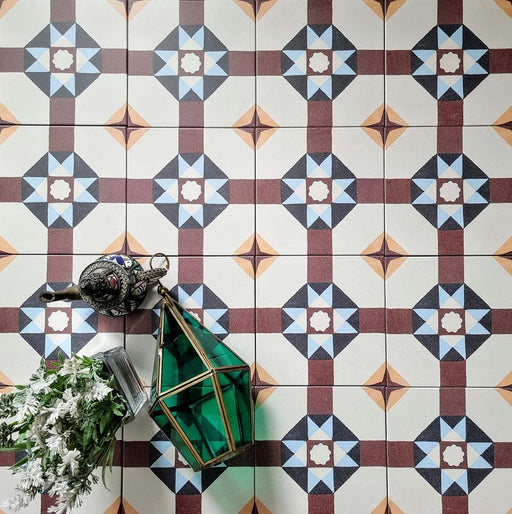 Overhead view of Europa Berlin 20x20cm Victorian Moroccan style floor tiles in burgundy, blue, mustard and cream tones, forming a geometric repeating pattern, styled with decorative lantern and floral arrangement.