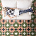 Marketing image featuring Geometry Moon 20x20cm geometric patterned floor tiles in green, terracotta, black, and cream tones, shown in a living room setting with a person relaxing on a white sofa while reading.
