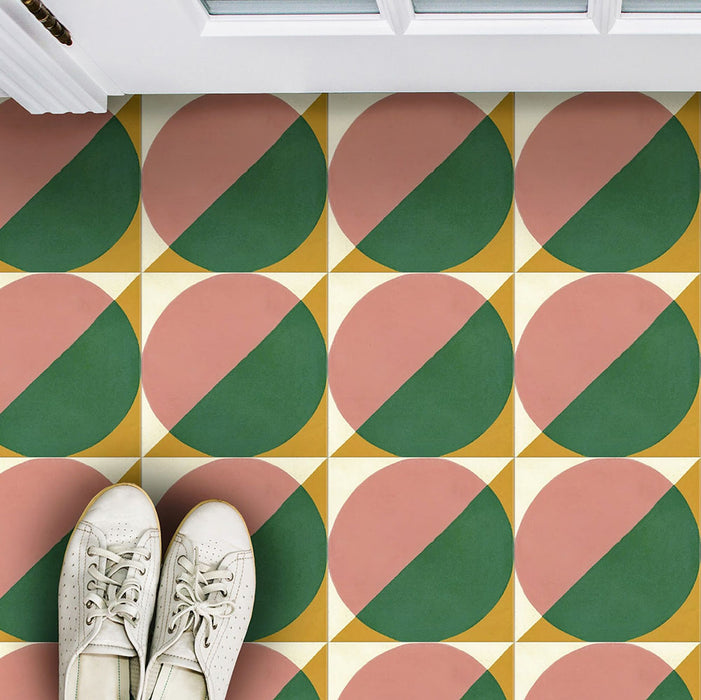 Marketing image featuring Geometry Paloma 20x20cm geometric patterned floor tiles in pink, green, mustard, and cream tones, shown from above with a pair of white trainers at a doorway.