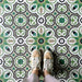 Overhead view of Europa Budapest 20x20cm Victorian Moroccan style floor tiles in green, cream and black tones, forming an intricate geometric pattern, styled with a person wearing trainers standing on the tiled floor.