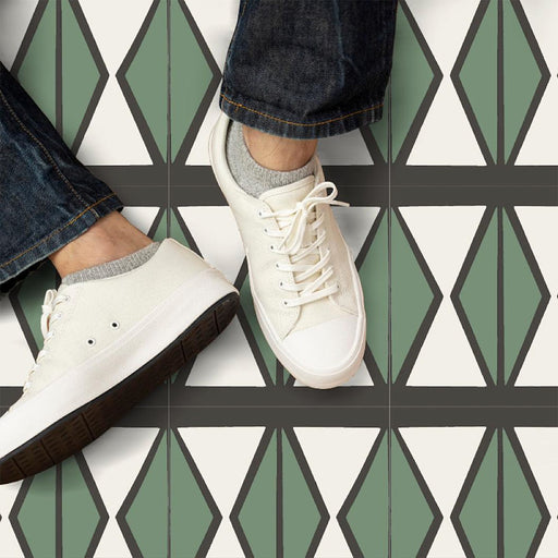 Marketing image featuring Geometry Hourglass 20x20cm patterned floor tiles with green diamond-shaped motifs and charcoal outlines on a cream background, shown from above with a person wearing white trainers and denim jeans standing on the tiles.
