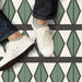 Marketing image featuring Geometry Hourglass 20x20cm patterned floor tiles with green diamond-shaped motifs and charcoal outlines on a cream background, shown from above with a person wearing white trainers and denim jeans standing on the tiles.
