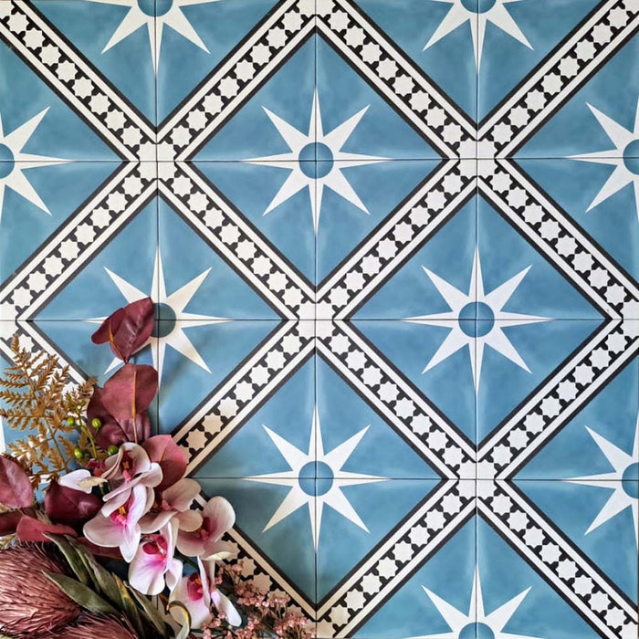 Galata Blue Moroccan Style 20x20cm tiles arranged in a star and diamond pattern featuring white starburst motifs and black geometric borders on a soft blue background, styled with pink and burgundy florals in the corner for a decorative marketing setting.