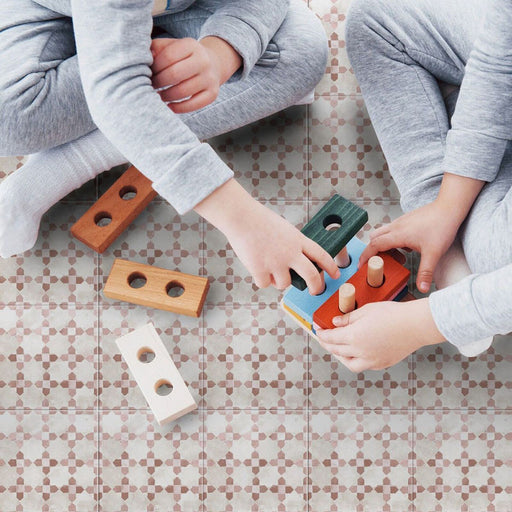 Bedouin Mequinez Rose Moroccan-style 15x15cm tiles used as flooring, featuring a soft blush pink and beige geometric pattern with subtle white detailing. Two children sit on the tiles playing with wooden toys, highlighting the tile’s warm, homely aesthetic and matte finish.