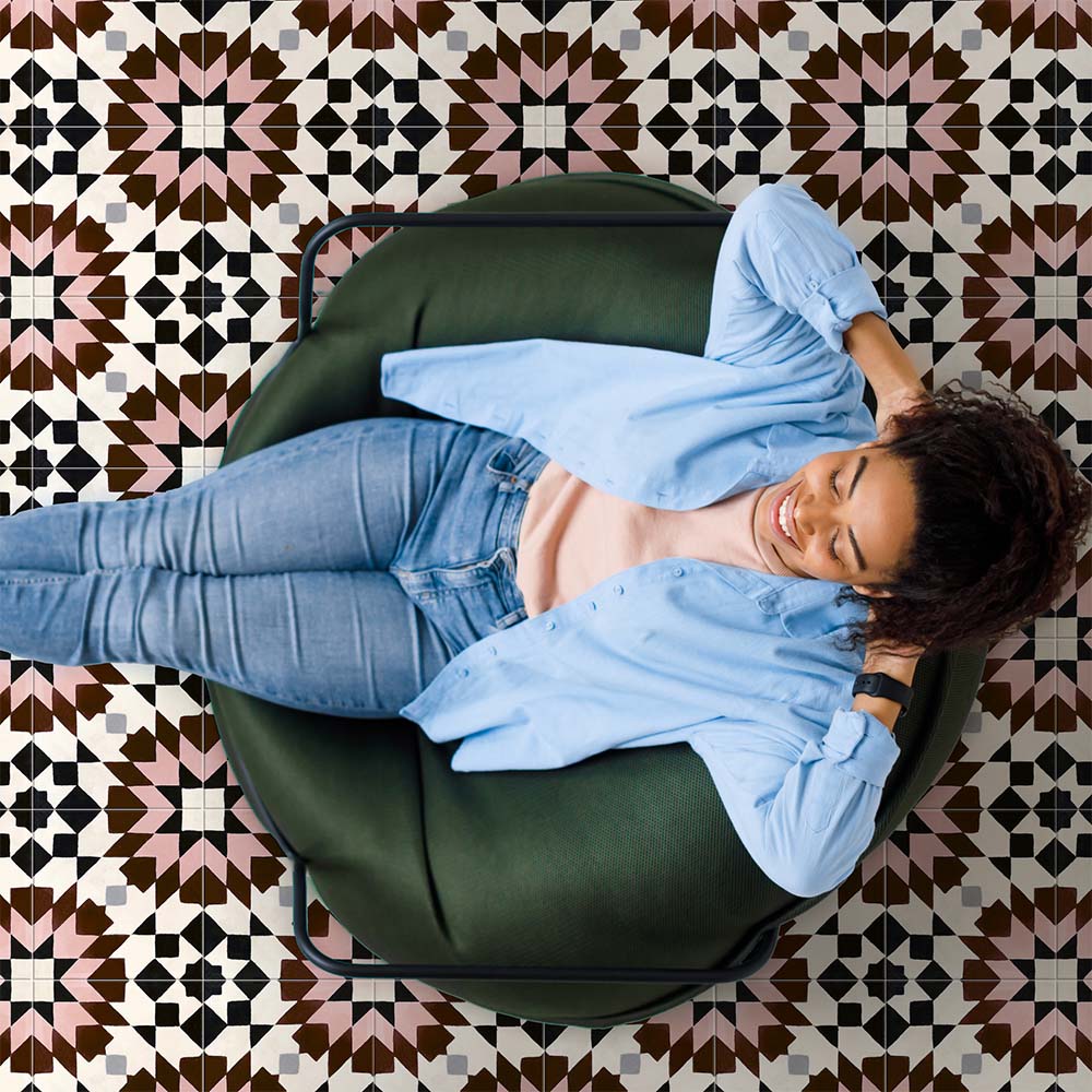 Cabana Mexico City Rose Moroccan Style 20x20cm tiles shown in a marketing setting with a person relaxing on a green chair. The floor features a geometric star pattern in rose pink, brown, black, and white with a smooth matt finish