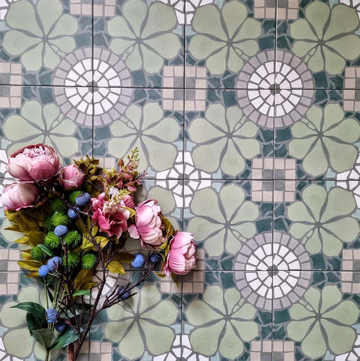 Bloom Green 20x20cm Moroccan-style ceramic tiles in a marketing flat-lay setting, featuring intricate floral patterns in green, white, and grey tones, styled with pink and blue flowers for a decorative touch.