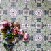 Bloom Green 20x20cm Moroccan-style ceramic tiles in a marketing flat-lay setting, featuring intricate floral patterns in green, white, and grey tones, styled with pink and blue flowers for a decorative touch.