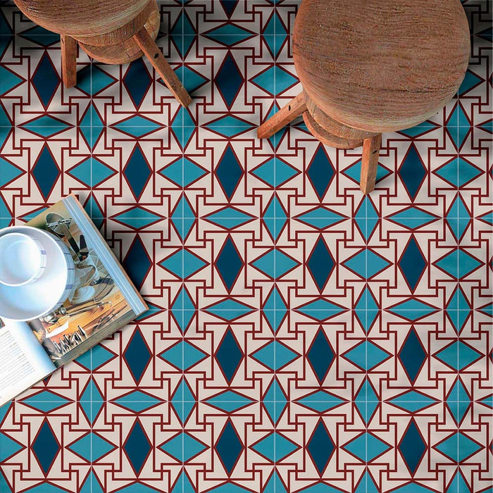Marketing image featuring Geometry Iris 20x20cm patterned floor tiles with an intricate geometric design in teal, navy, and terracotta-red tones on a cream base, shown beneath wooden stools and an open magazine to highlight the bold repeating pattern.