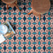 Marketing image featuring Geometry Iris 20x20cm patterned floor tiles with an intricate geometric design in teal, navy, and terracotta-red tones on a cream base, shown beneath wooden stools and an open magazine to highlight the bold repeating pattern.