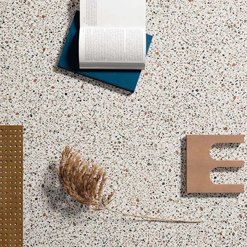 Flatlay featuring Dino Terrazzo Blanco Dots 20x20cm terrazzo-style ceramic tiles, styled with an open book, dried foliage, and decorative accessories on a speckled white surface.