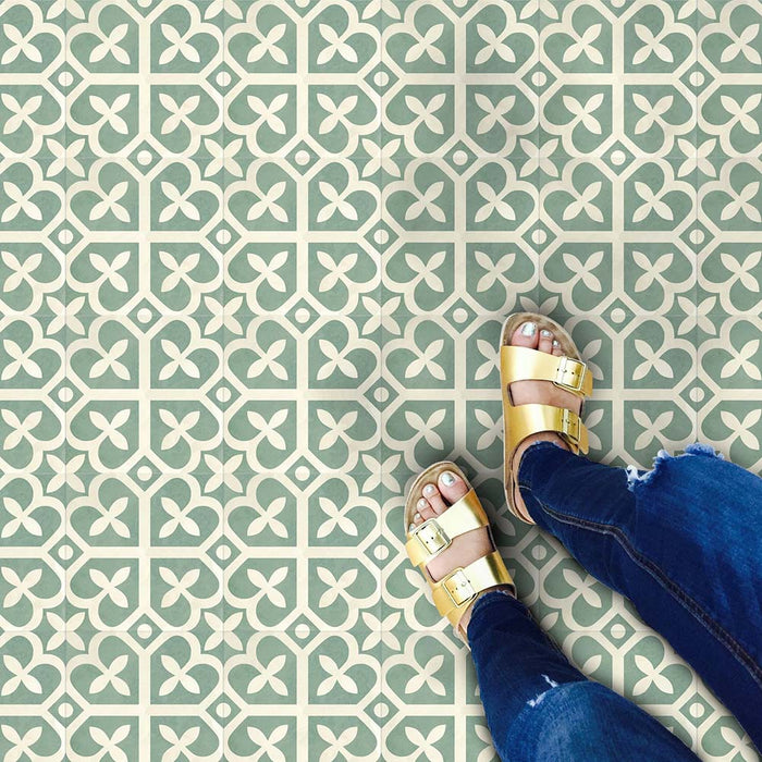 Marketing setting featuring Iris Green Victorian Moroccan-style 20×20cm porcelain floor tiles, showcasing a soft green and cream geometric pattern styled with casual denim and gold sandals for a playful, lifestyle-led interior scene.